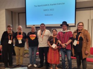 Facilitators of the World KAIROS Blanket Exercise stand together under a presentation that reads: The World KAIROS Blanket Exercise. WIPCE 2022. Welcome.